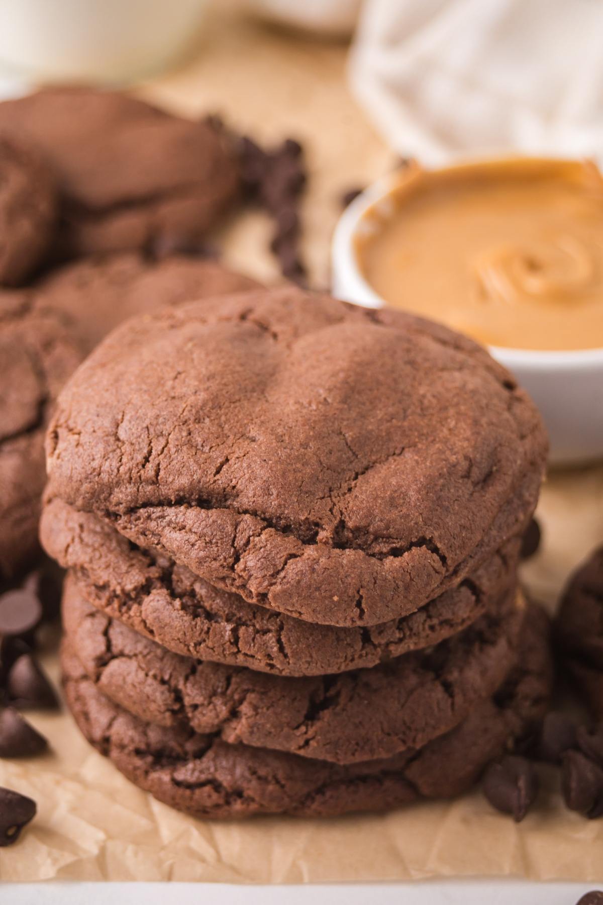 A stack of three chocolate cookies next to chocolate chips and a bowl of peanut butter.