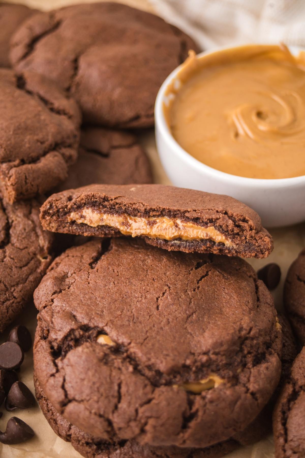A chocolate cookie broken in half reveals a peanut butter filling, with more cookies and a bowl of peanut butter nearby.