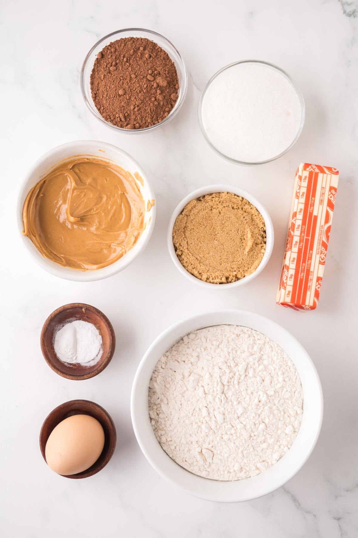 Baking ingredients in bowls: flour, cocoa, sugars, peanut butter, butter stick, egg, salt, and baking soda.