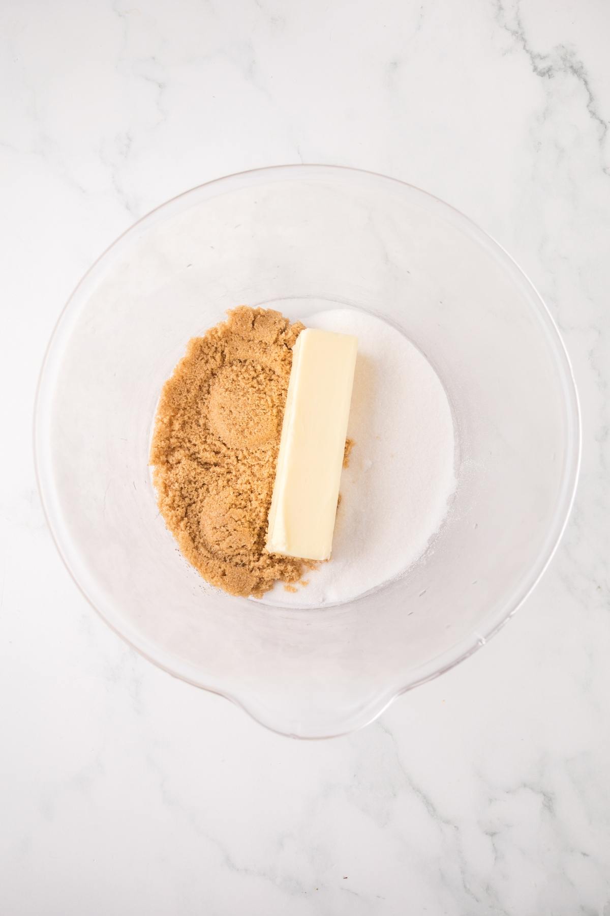 A mixing bowl with a stick of butter, brown sugar, and white sugar on a marble surface.