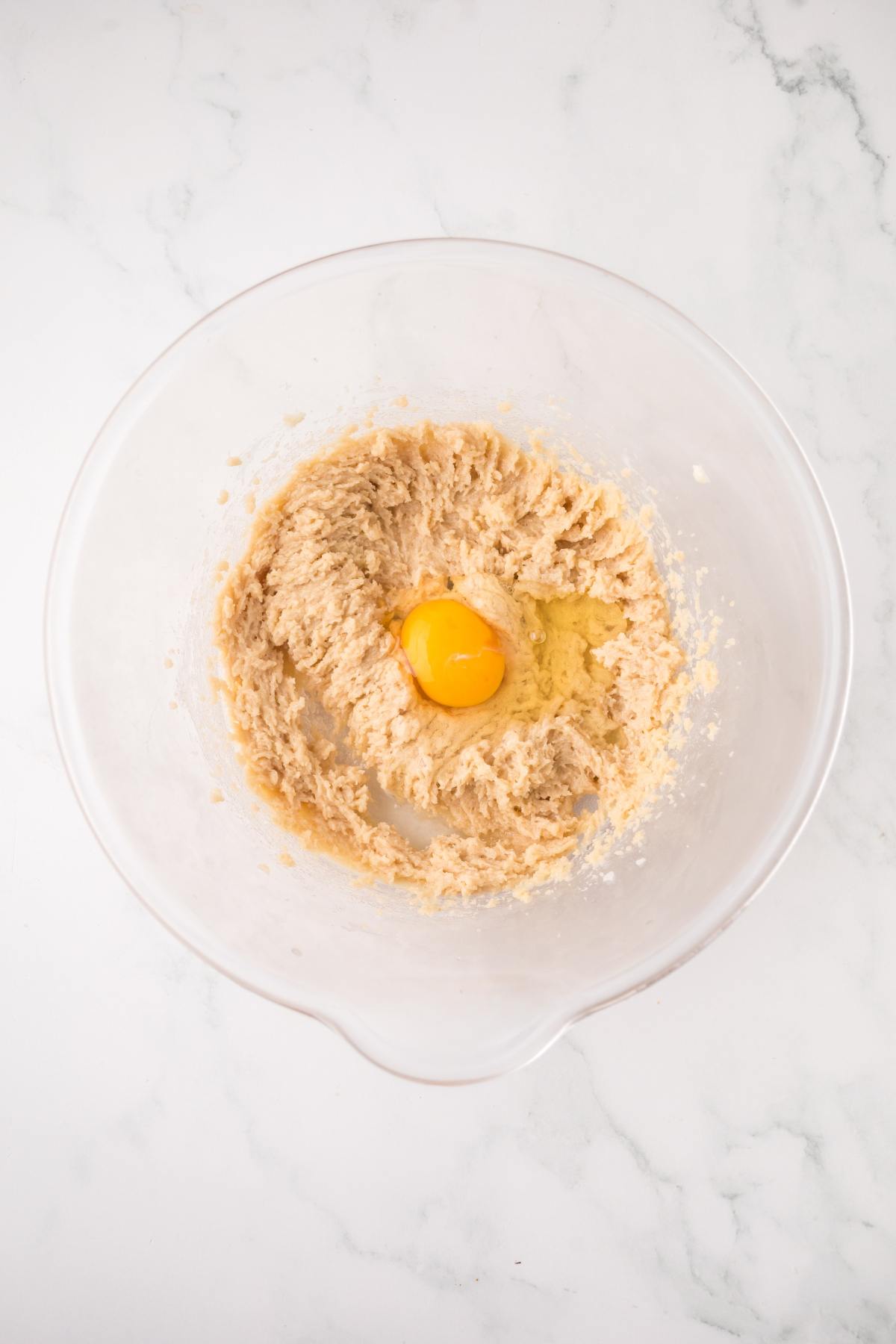 A mixing bowl with creamed dough and a raw egg in the center, on a white marble surface.