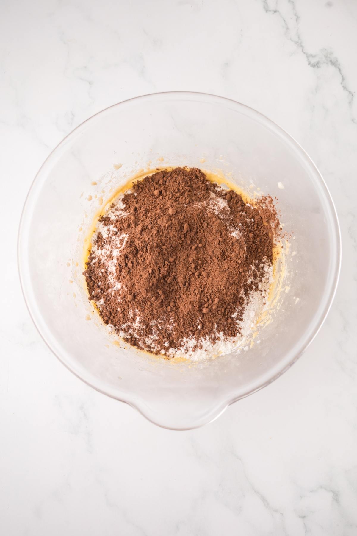 Clear glass bowl with flour and cocoa powder mixture on a white marble surface.