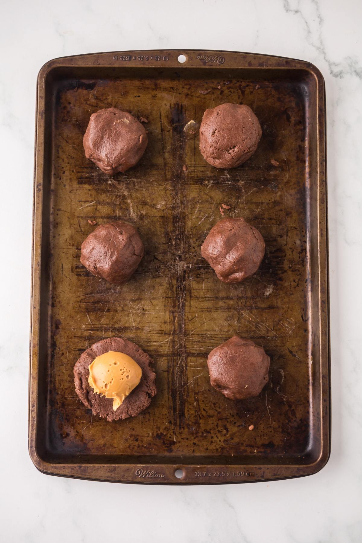 Six chocolate cookie dough balls on a baking sheet, one with a piece of peanut butter inside.