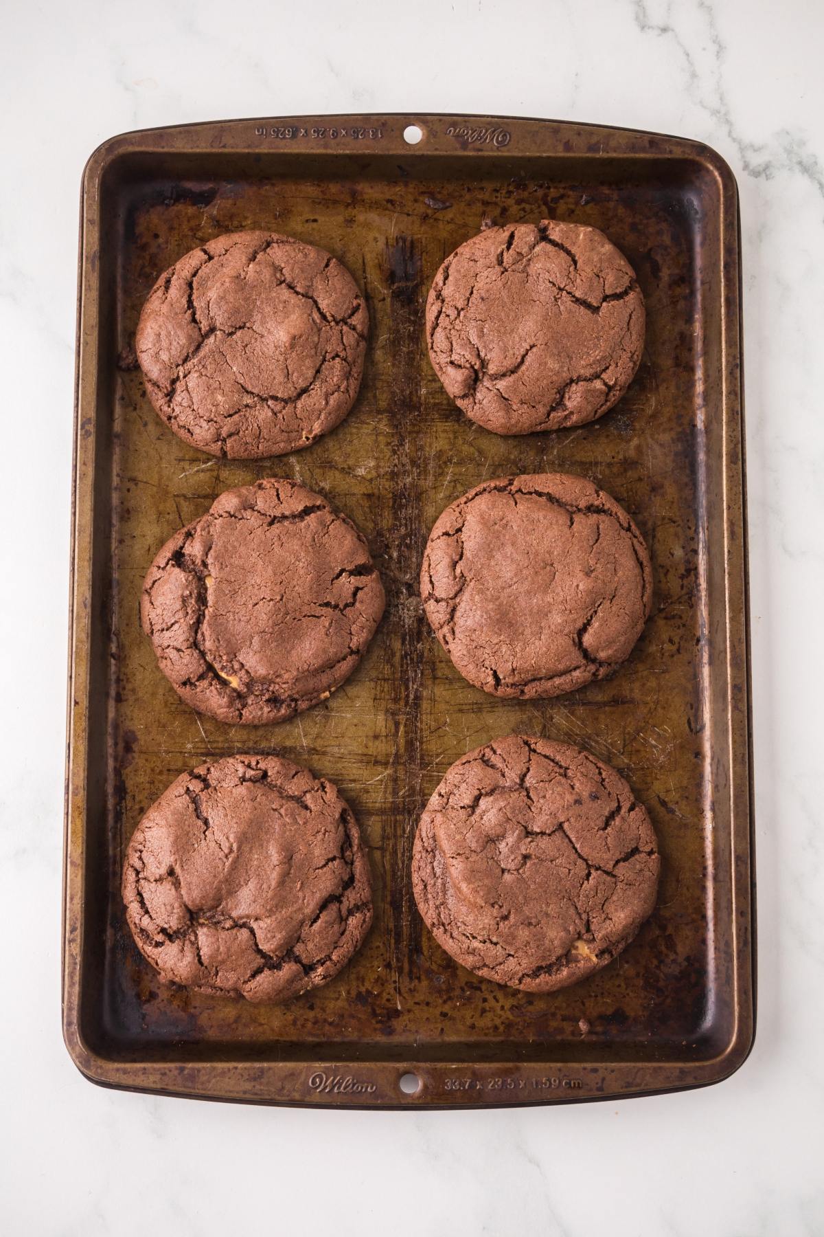 Six large chocolate cookies on a worn baking sheet, placed on a white marble surface.