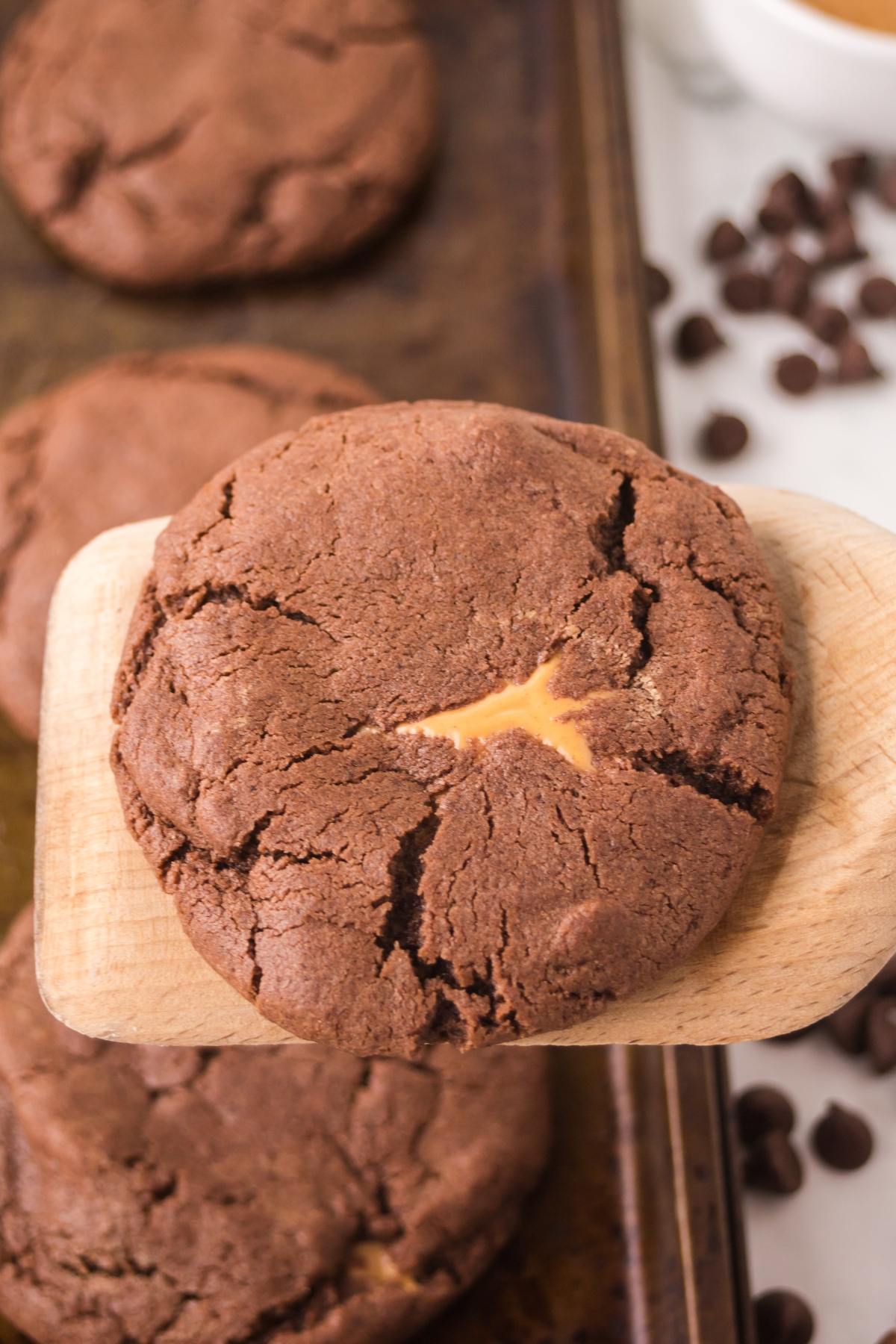 A chocolate cookie with a caramel center on a wooden spatula, with more cookies and chocolate chips in the background.