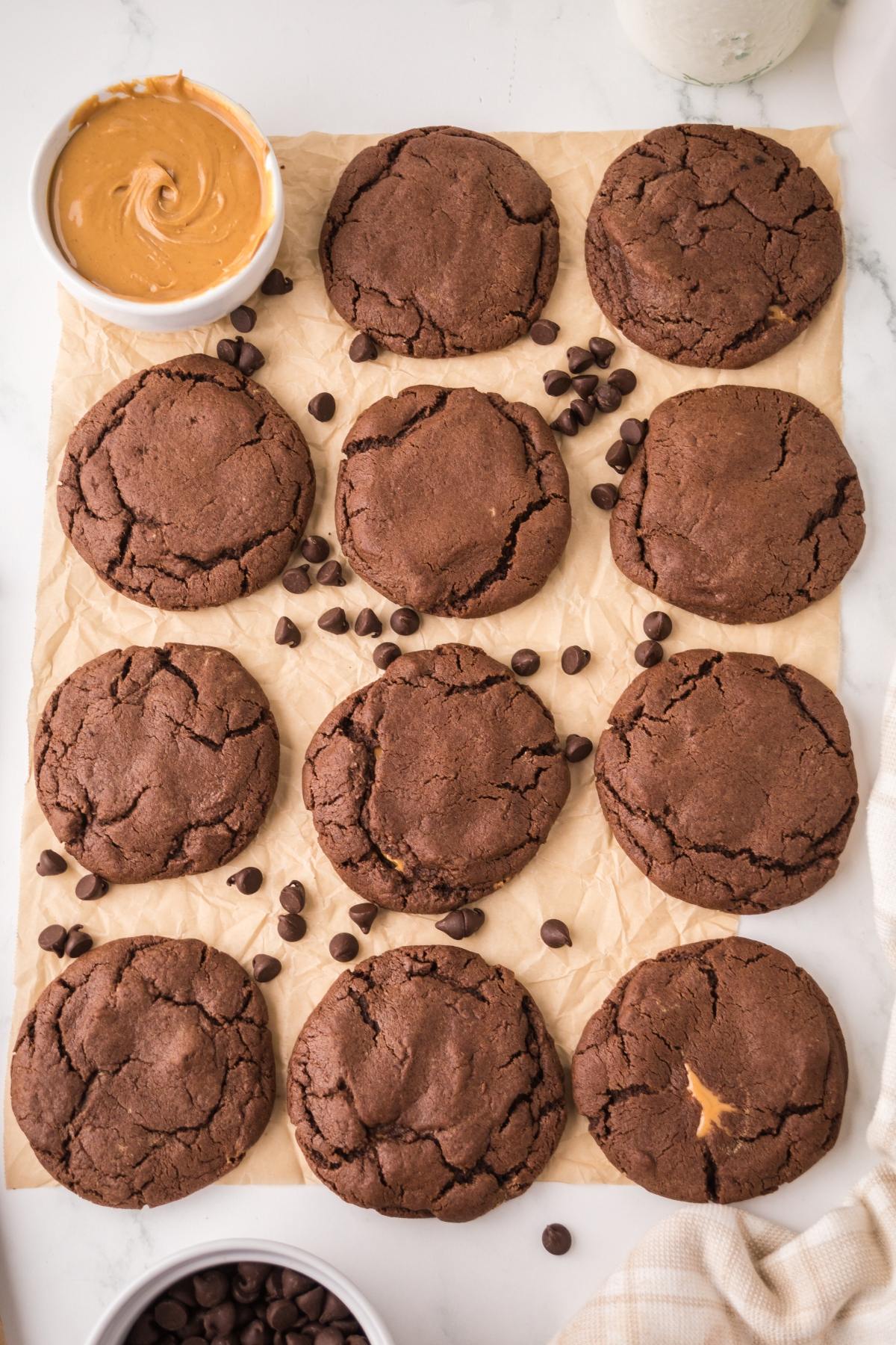 Twelve chocolate cookies on parchment with scattered chocolate chips and a bowl of peanut butter nearby.