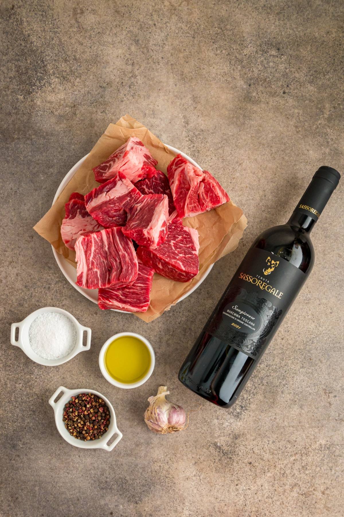 Raw beef chunks on a plate, a bottle of red wine, salt, peppercorns, olive oil, and garlic on a table.