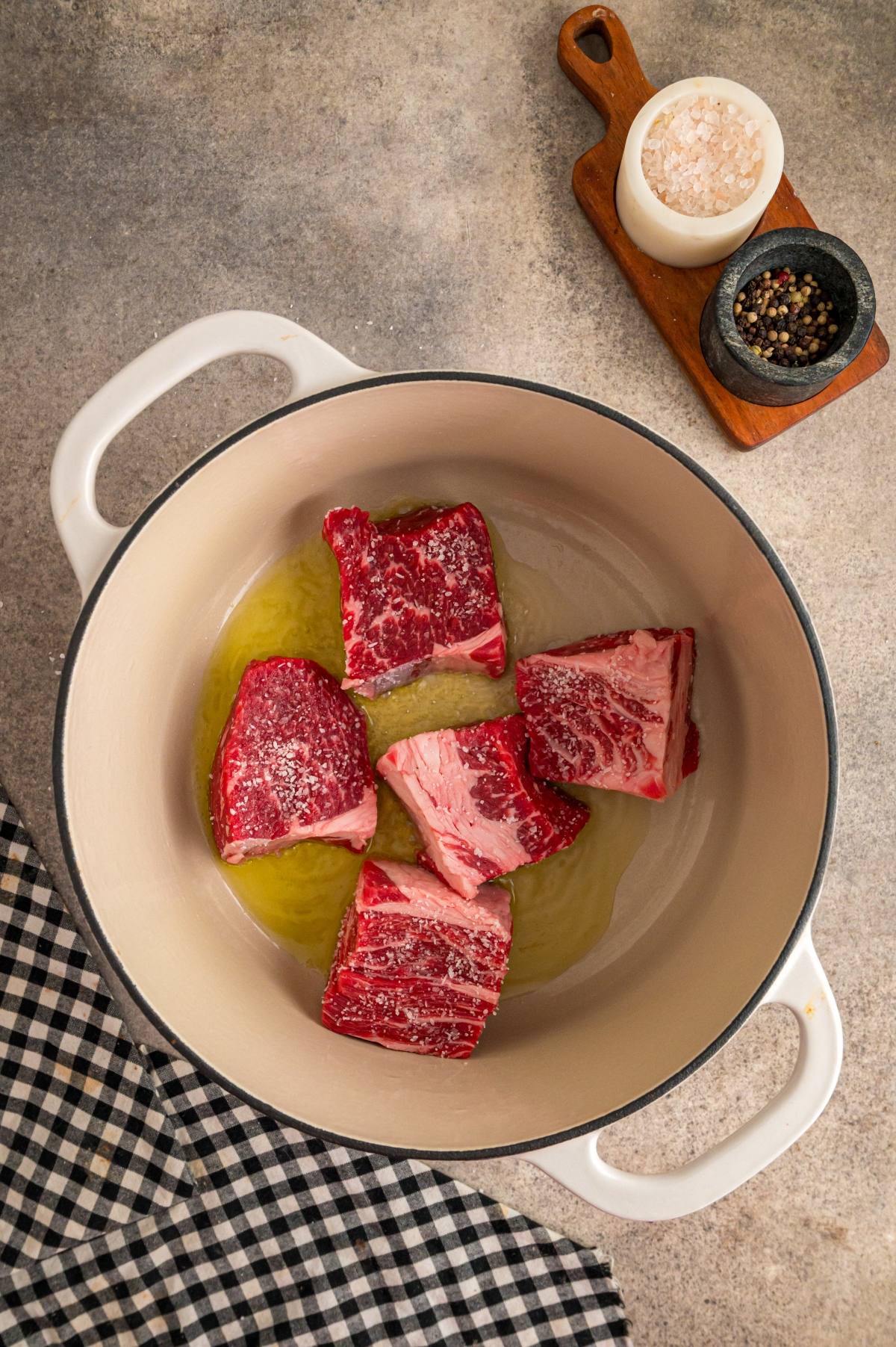Four raw beef short ribs in a white pot with oil, next to salt, pepper, and a checkered cloth.