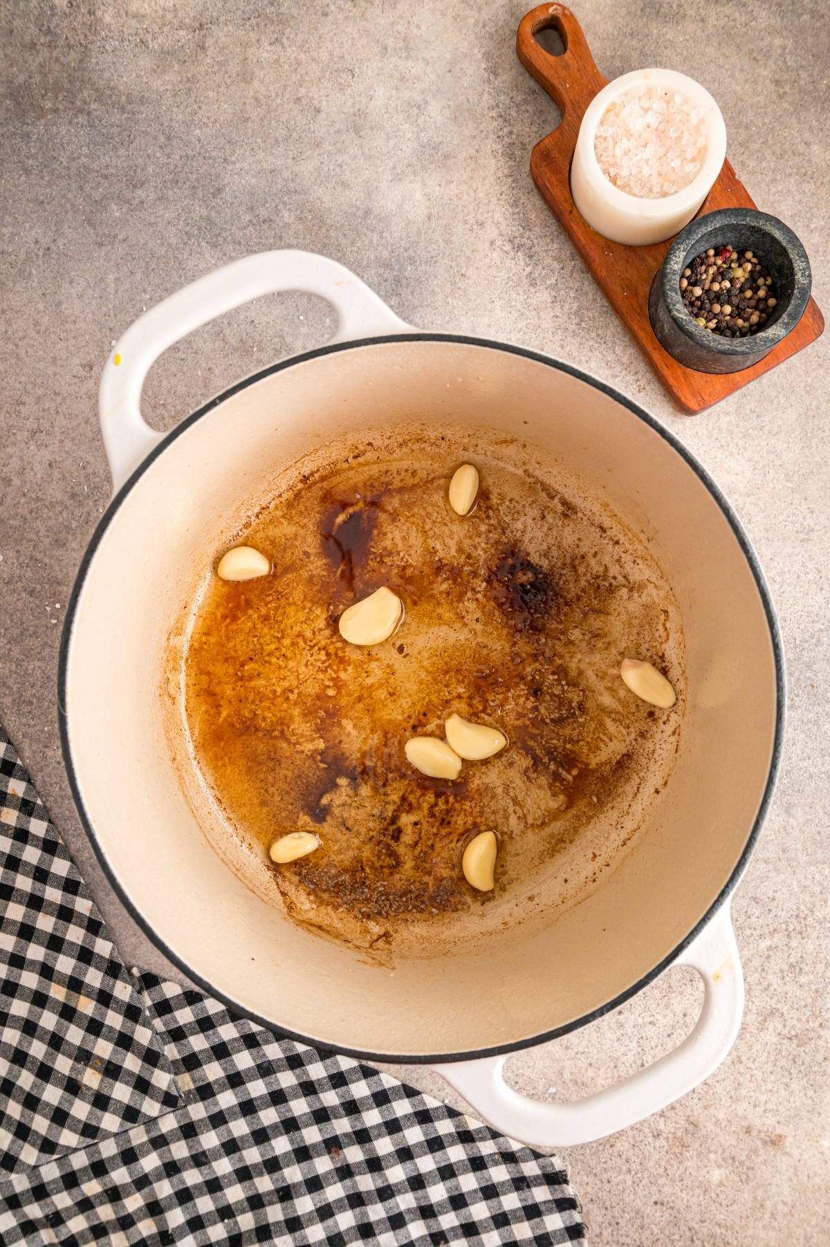 A white pot with browned bits and garlic cloves, next to salt, pepper, and a black-and-white checkered towel.