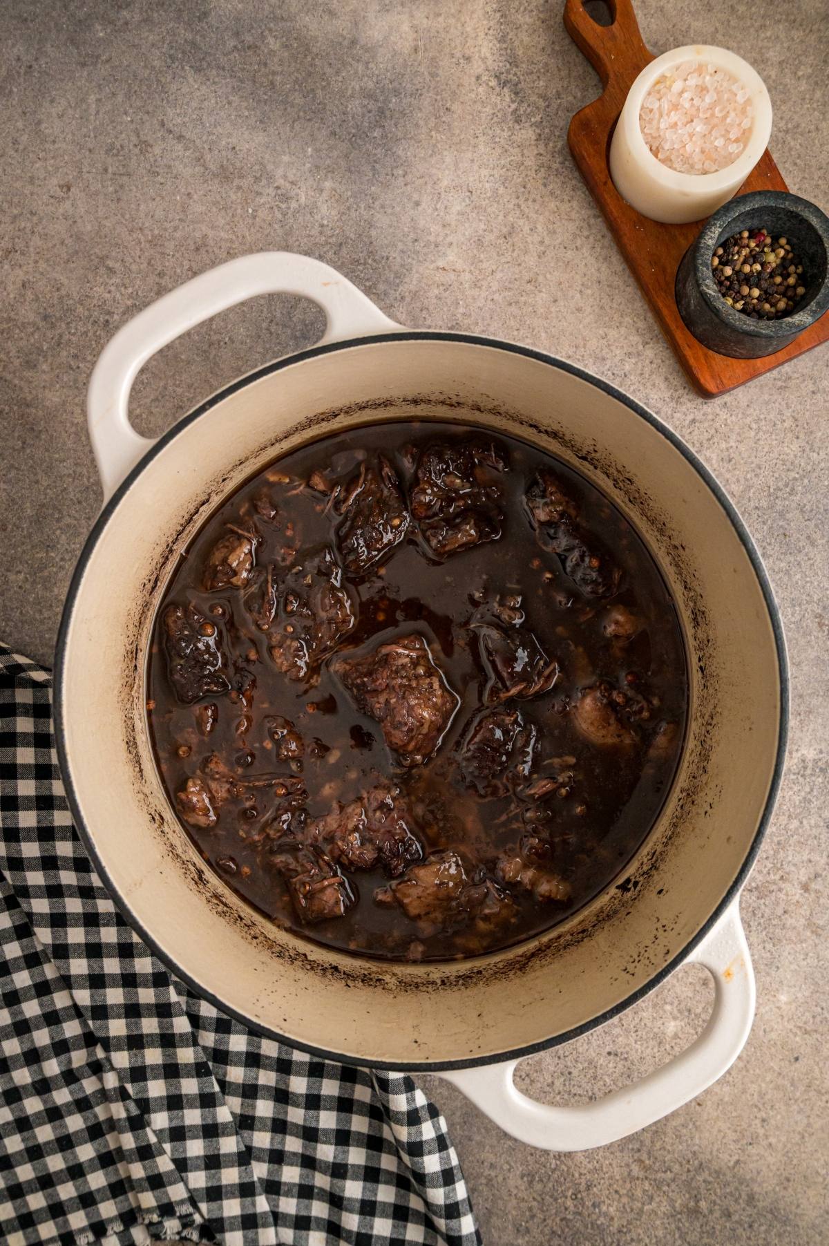 A white pot filled with braised beef stew, next to a checkered cloth, salt, and pepper on a gray surface.