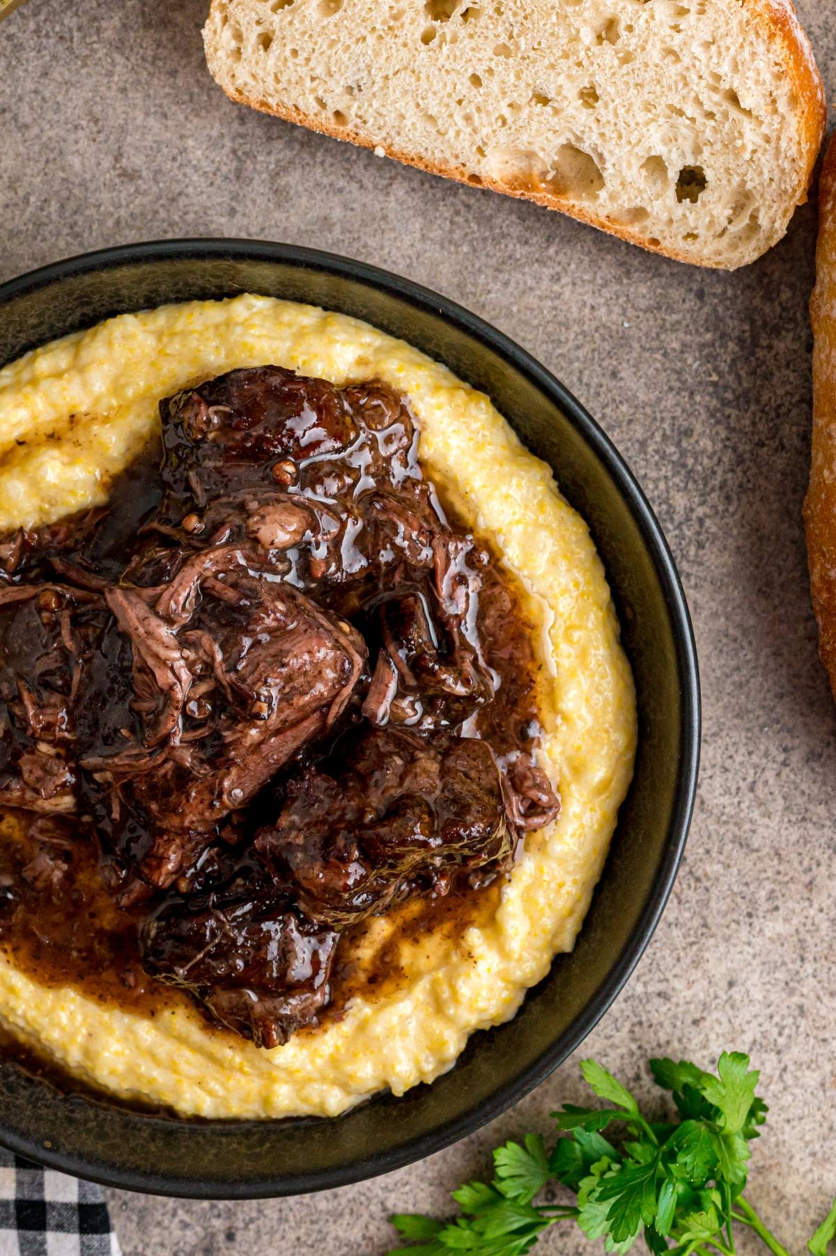 A bowl of creamy polenta topped with braised beef, served with slices of bread on the side.