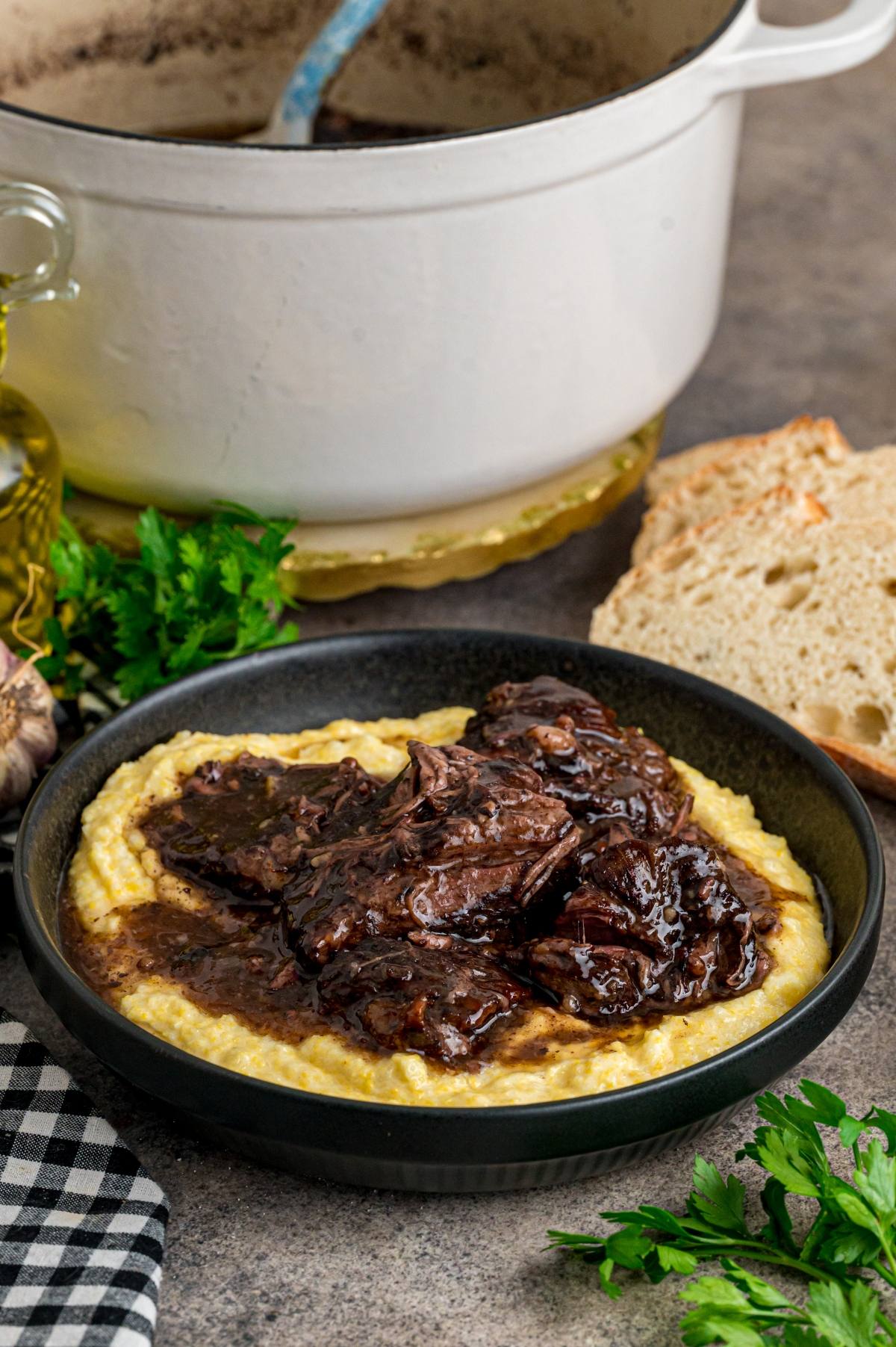 A bowl of creamy polenta topped with braised beef stew, beside bread, parsley, and a white pot.