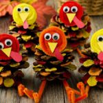 Five pine cone turkeys made from googly eyes, pipe cleaners, and colored paper are displayed on a wooden table.