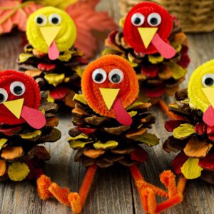Five pine cone turkeys made from googly eyes, pipe cleaners, and colored paper are displayed on a wooden table.