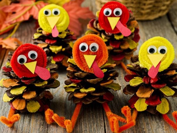 Five pine cone turkeys made from googly eyes, pipe cleaners, and colored paper are displayed on a wooden table.