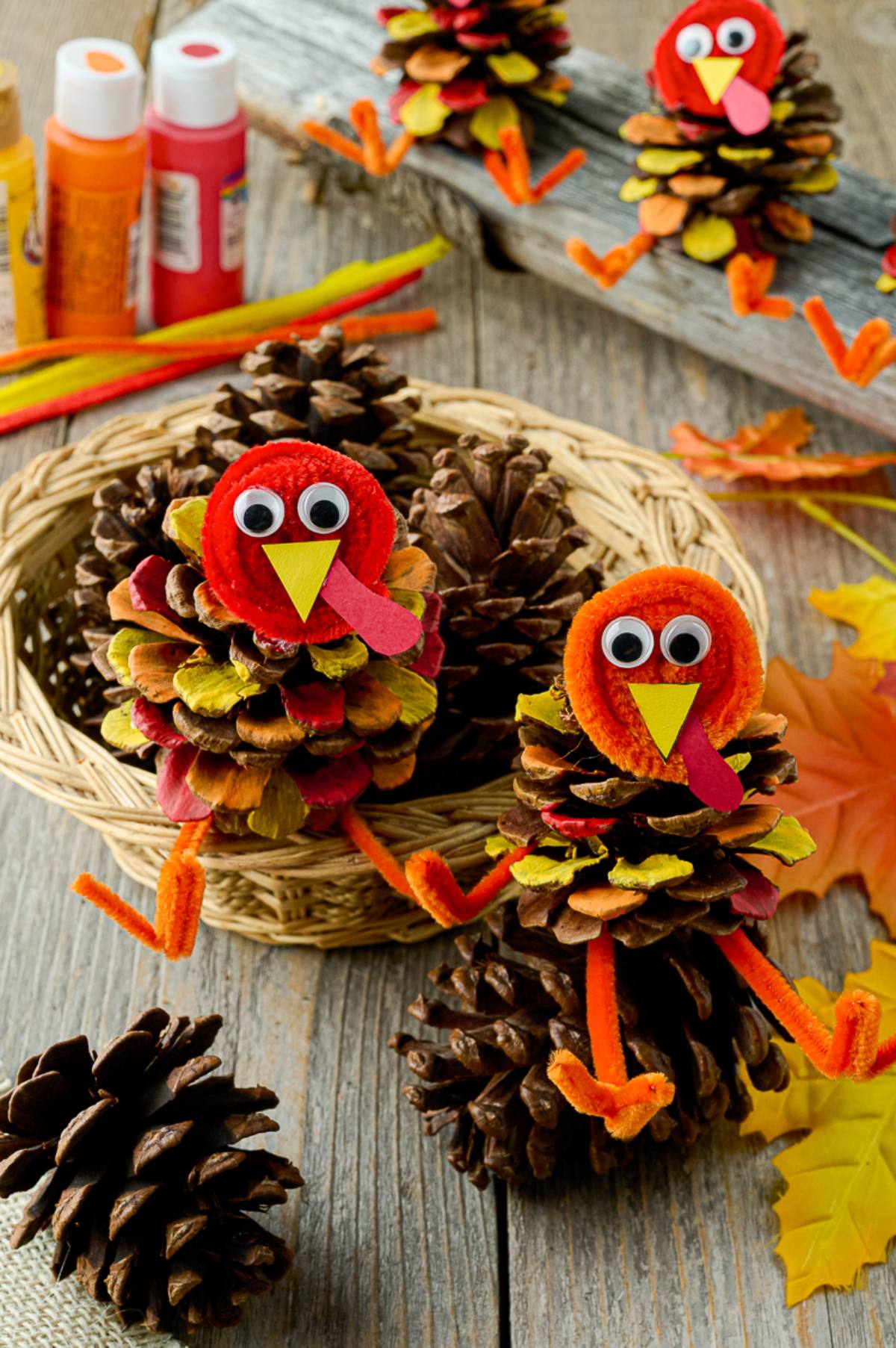 Pinecones decorated as turkeys with pipe cleaners and googly eyes, surrounded by autumn leaves and craft supplies.