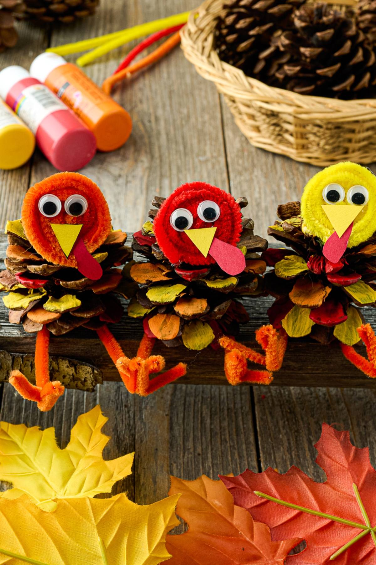Three turkey crafts made from pinecones, pipe cleaners, and googly eyes sit on a wooden surface with autumn leaves.