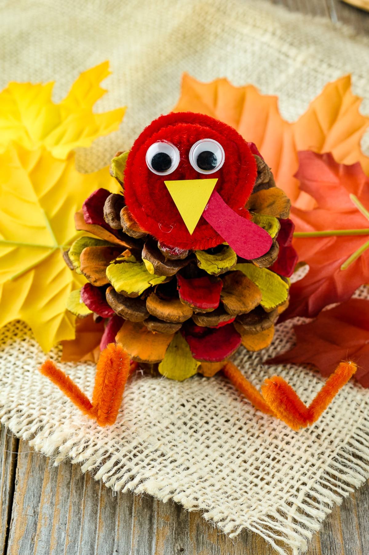 A pine cone turkey craft with googly eyes and pipe cleaner legs, on a burlap cloth with autumn leaves.
