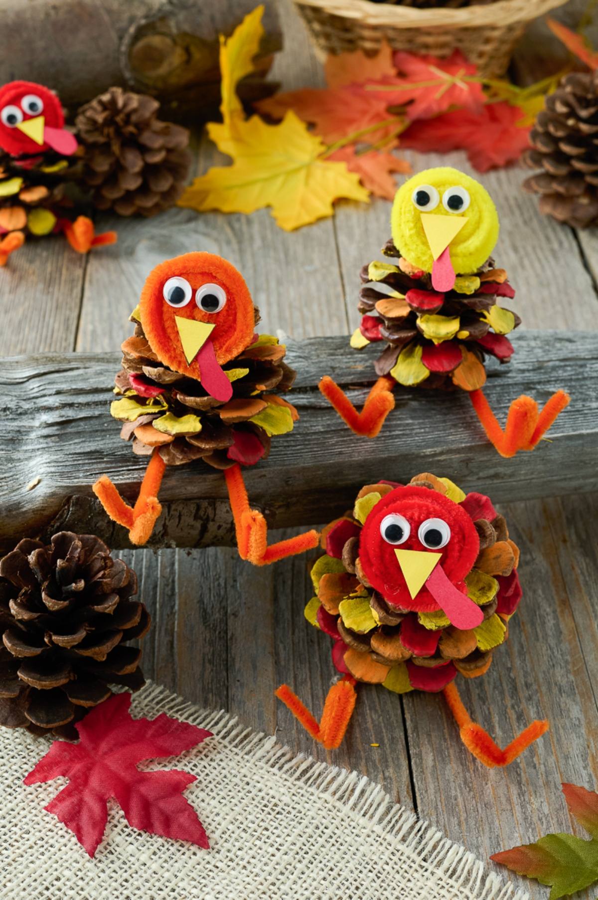 Three pinecone turkeys with googly eyes sit on a log, surrounded by pinecones and colorful autumn leaves.