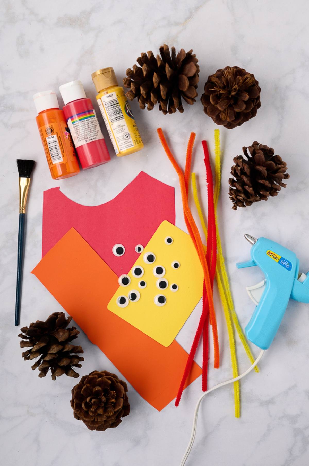 Art supplies, pinecones, paint, googly eyes, pipe cleaners, glue gun, and colored paper on a white surface.
