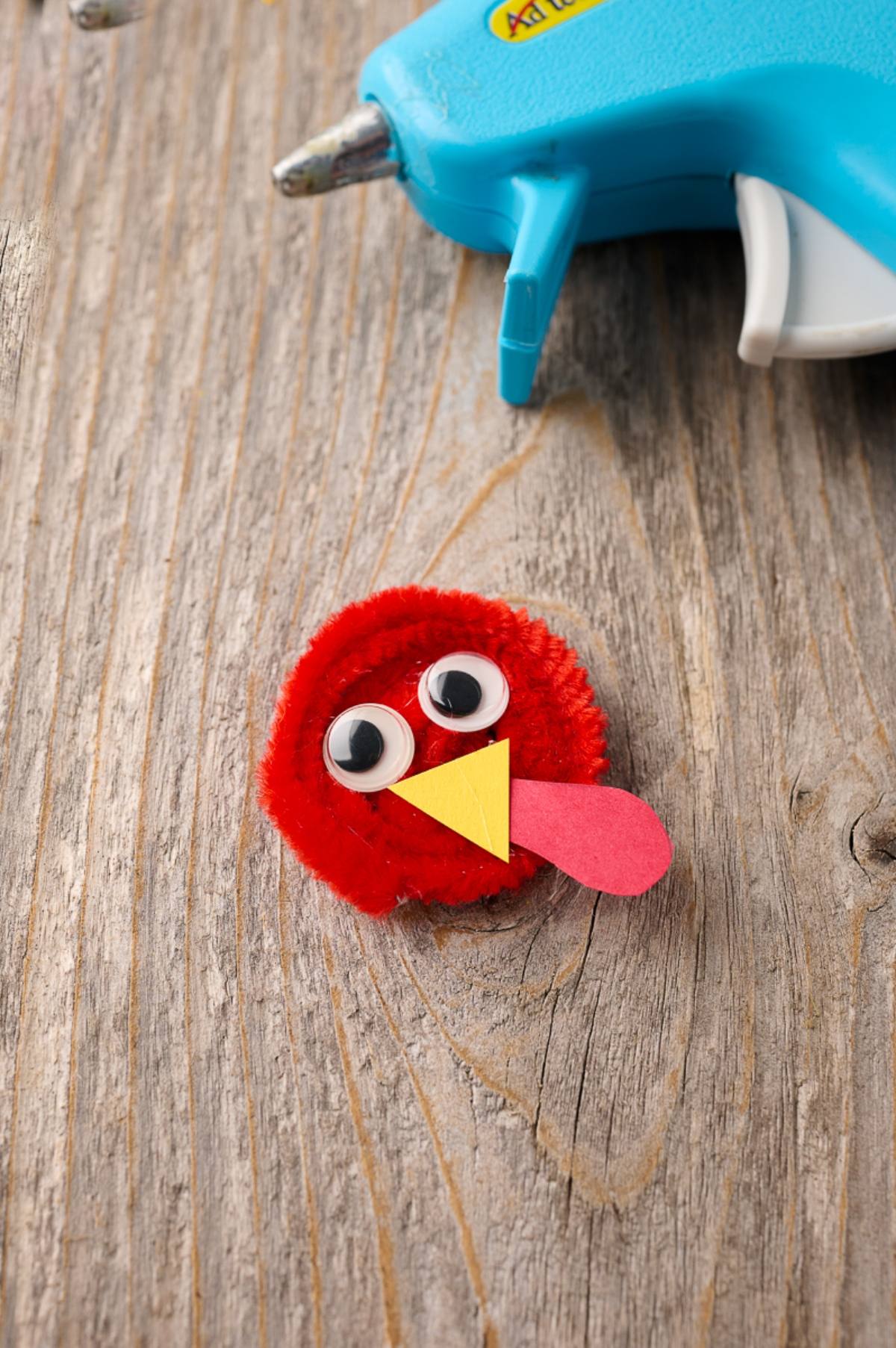 A red pom-pom turkey craft with googly eyes and paper beak, next to a blue hot glue gun on wood.