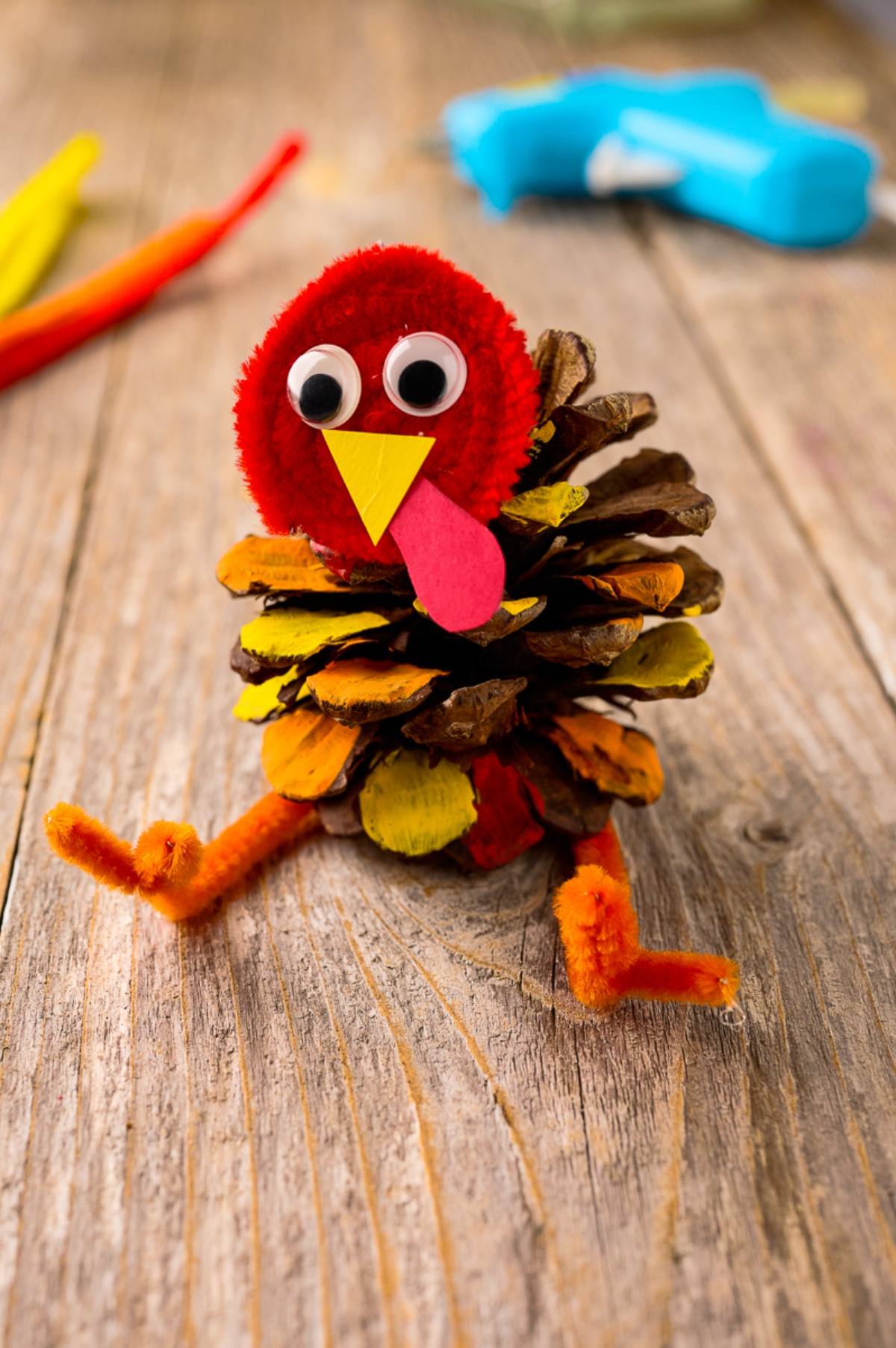 A turkey craft made from a pinecone, pipe cleaners, googly eyes, and colorful painted details on a wooden surface.