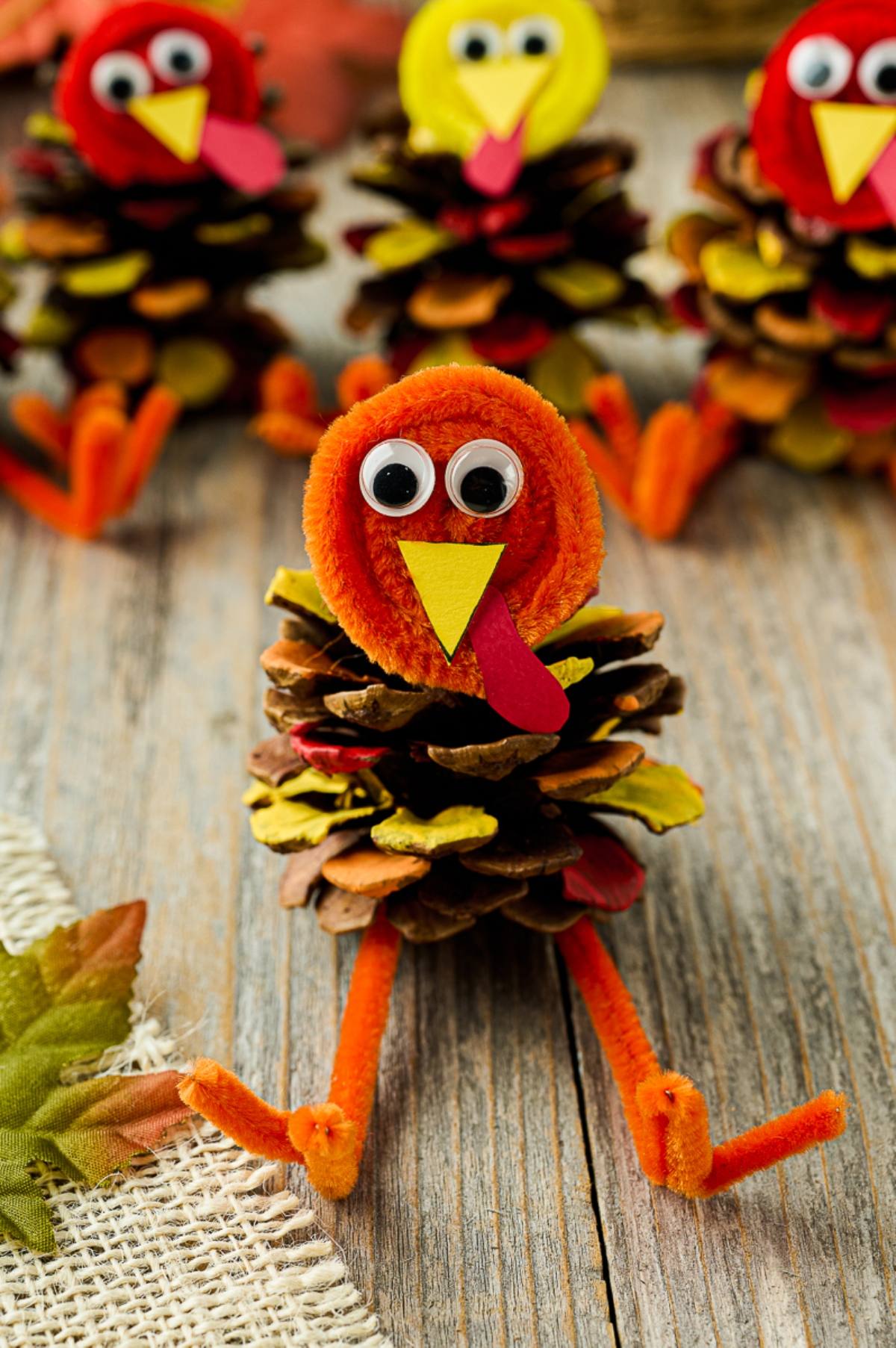 Pine cone turkey crafts with googly eyes and pipe cleaner beaks and legs on a wooden surface.