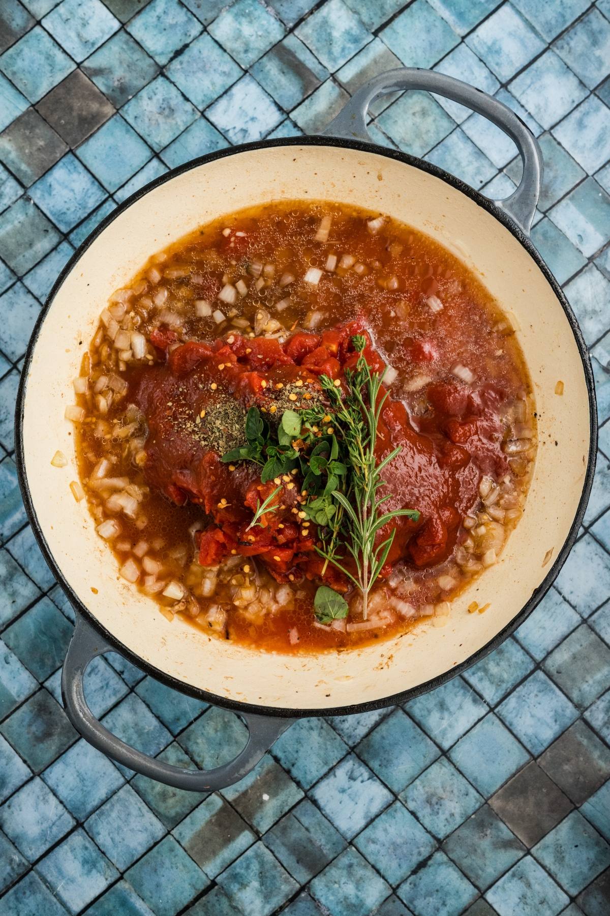 A pot of tomato sauce with onions, herbs, and seasonings on a blue tiled surface.