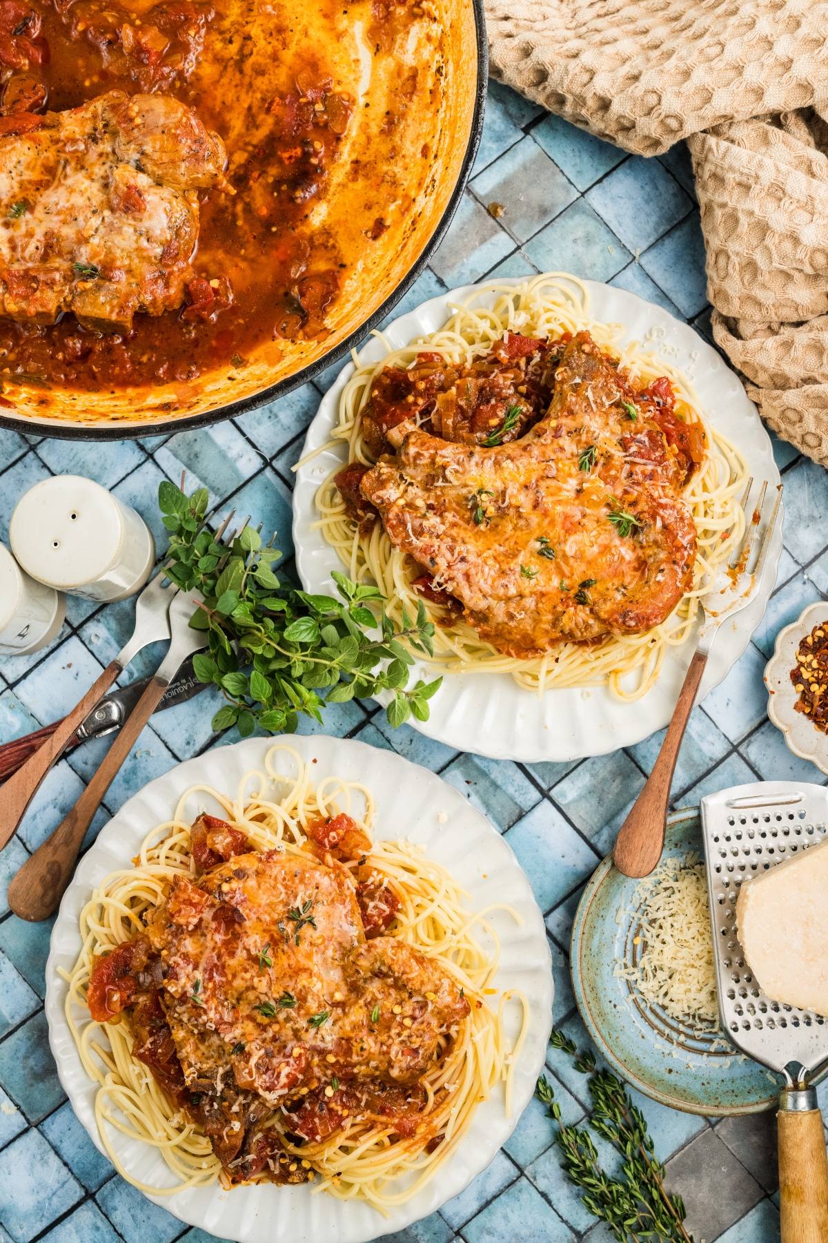 Two plates of spaghetti topped with saucy pork chops and herbs, with cheese and sauce pot nearby on a blue tiled table.