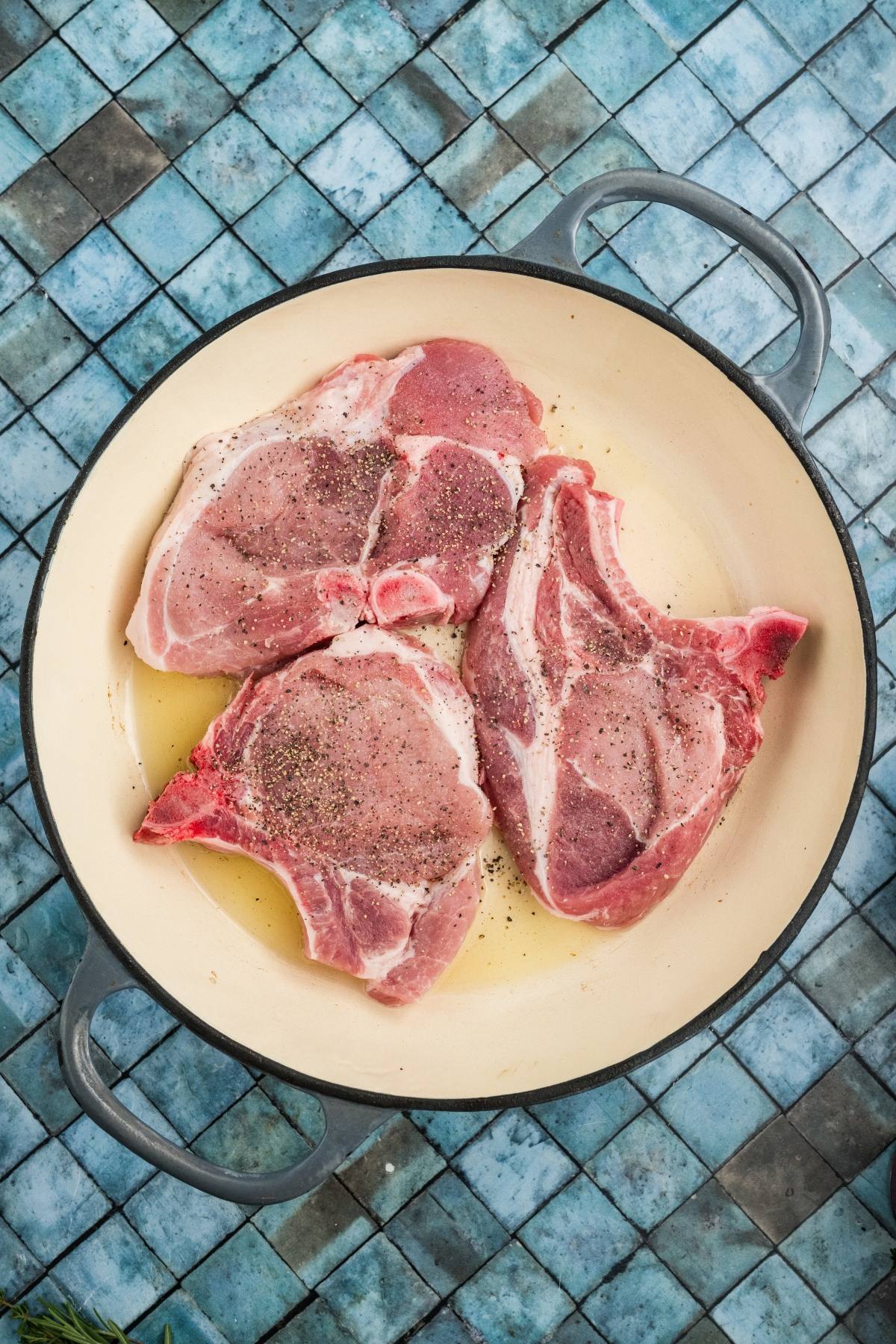 Three raw, bone-in pork chops seasoned with pepper in a round white pan on a blue tiled surface.