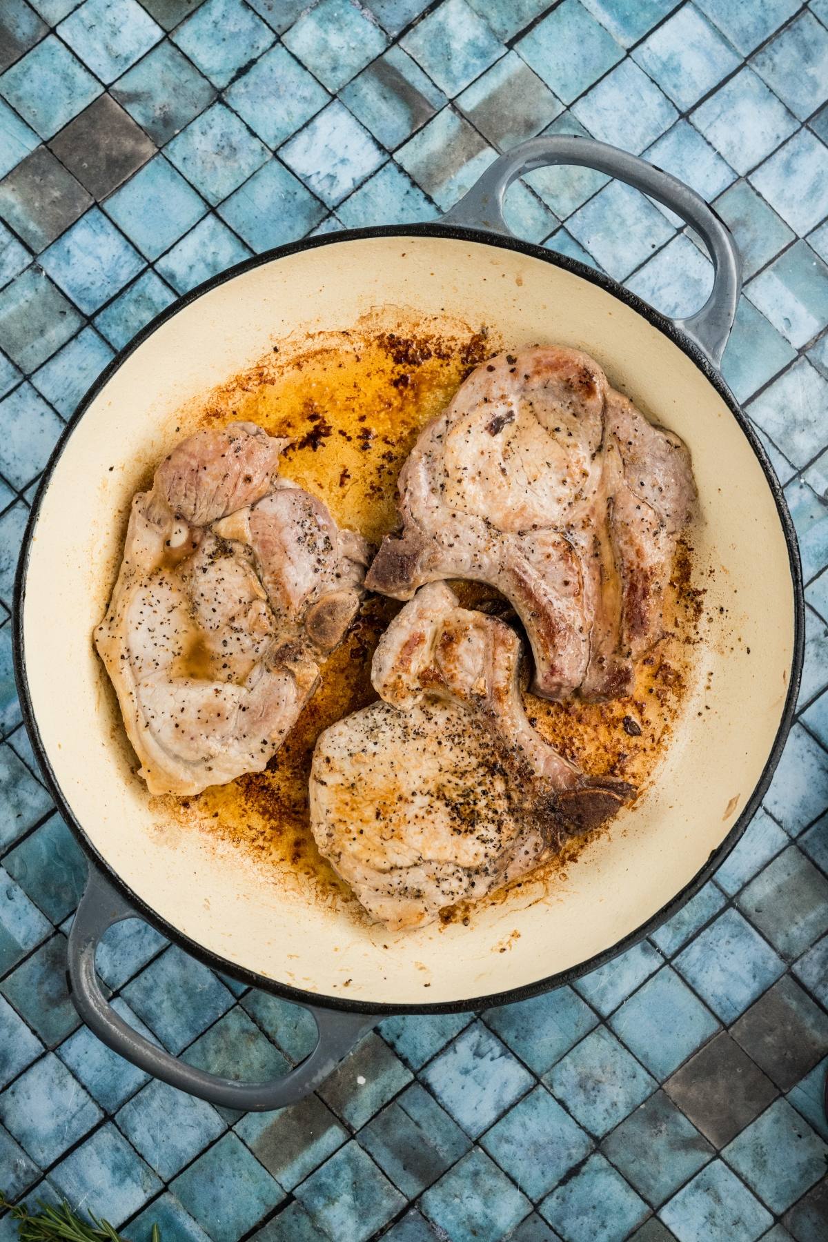Three browned pork chops in a white skillet on a blue tiled surface.