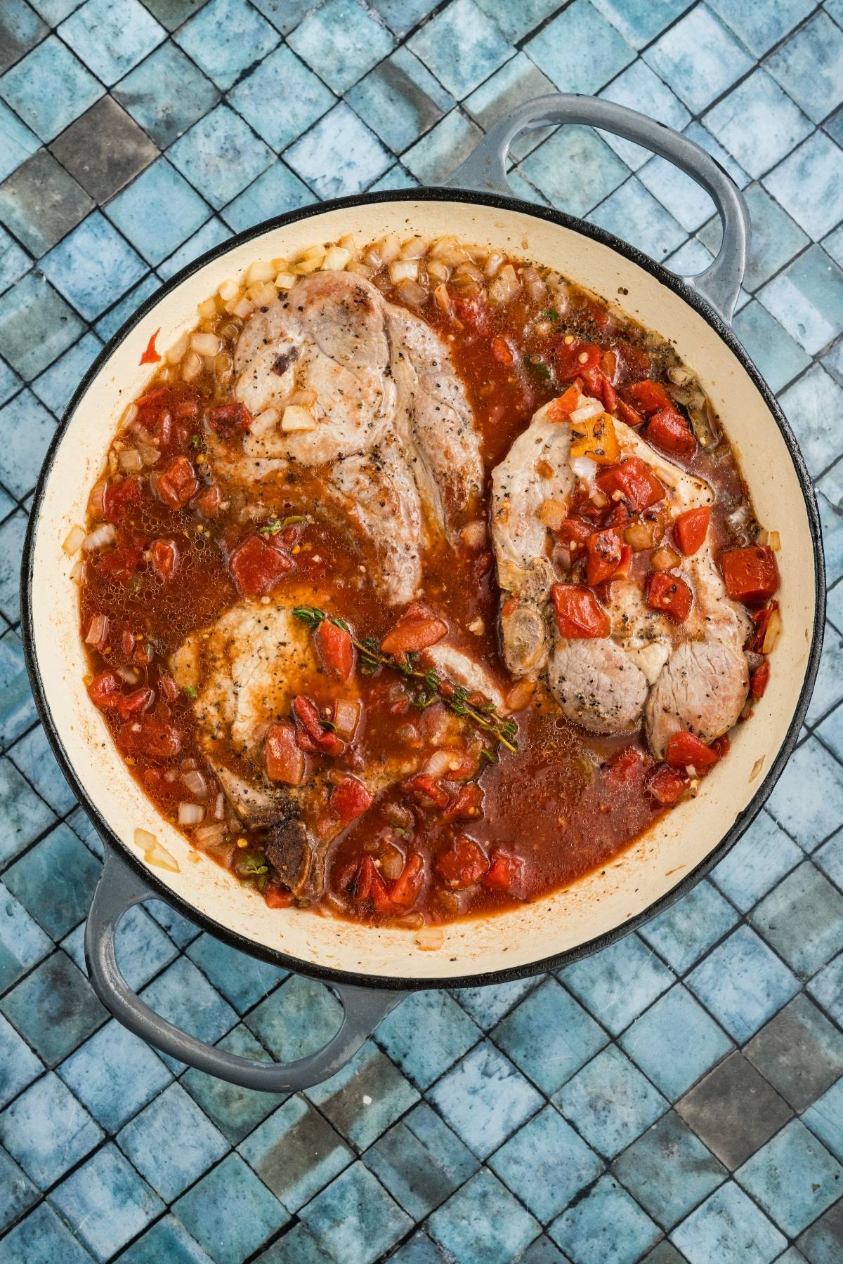 Chicken braised in tomato sauce with onions and herbs in a round pot on a blue-tiled surface.