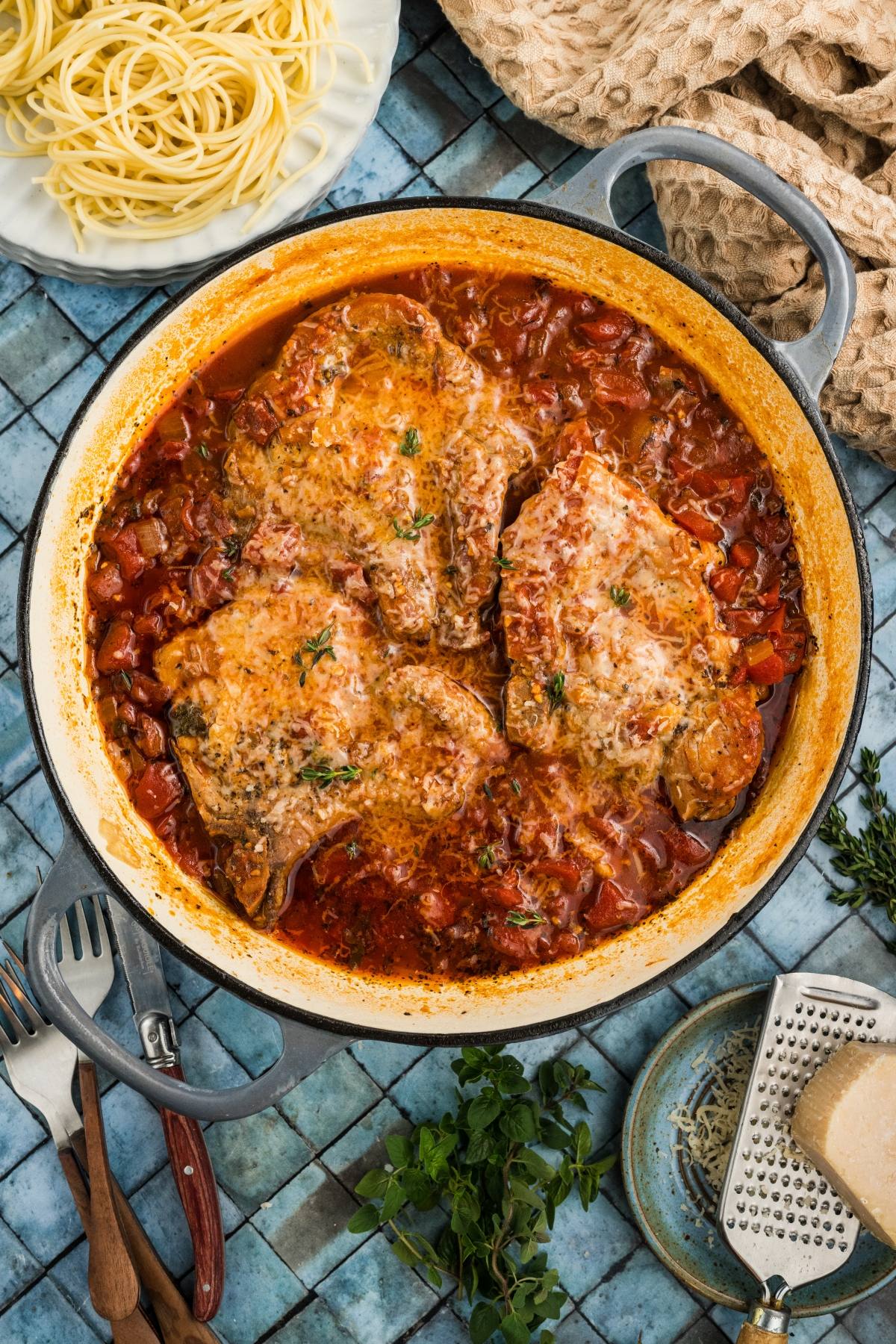 Braised meat in tomato sauce in a pot, with herbs, spaghetti, cheese, and utensils on a blue tiled table.