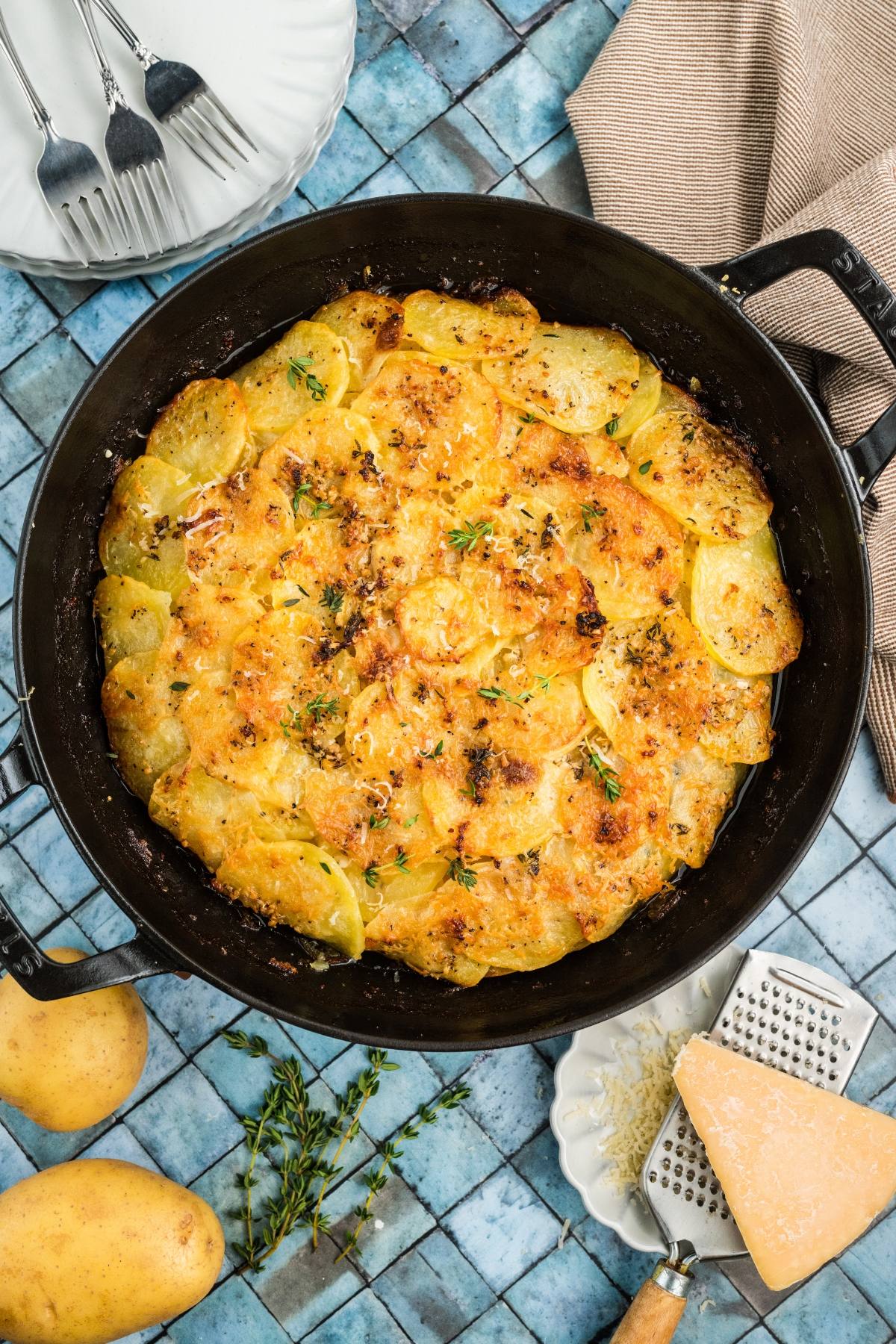 Golden baked potatoes galette in a black skillet, garnished with herbs, on a blue tile background.