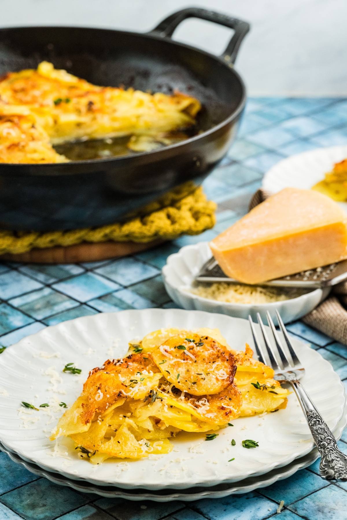 A plate of cheesy potato galette with herbs, with more gratin in a skillet and a wedge of cheese nearby.