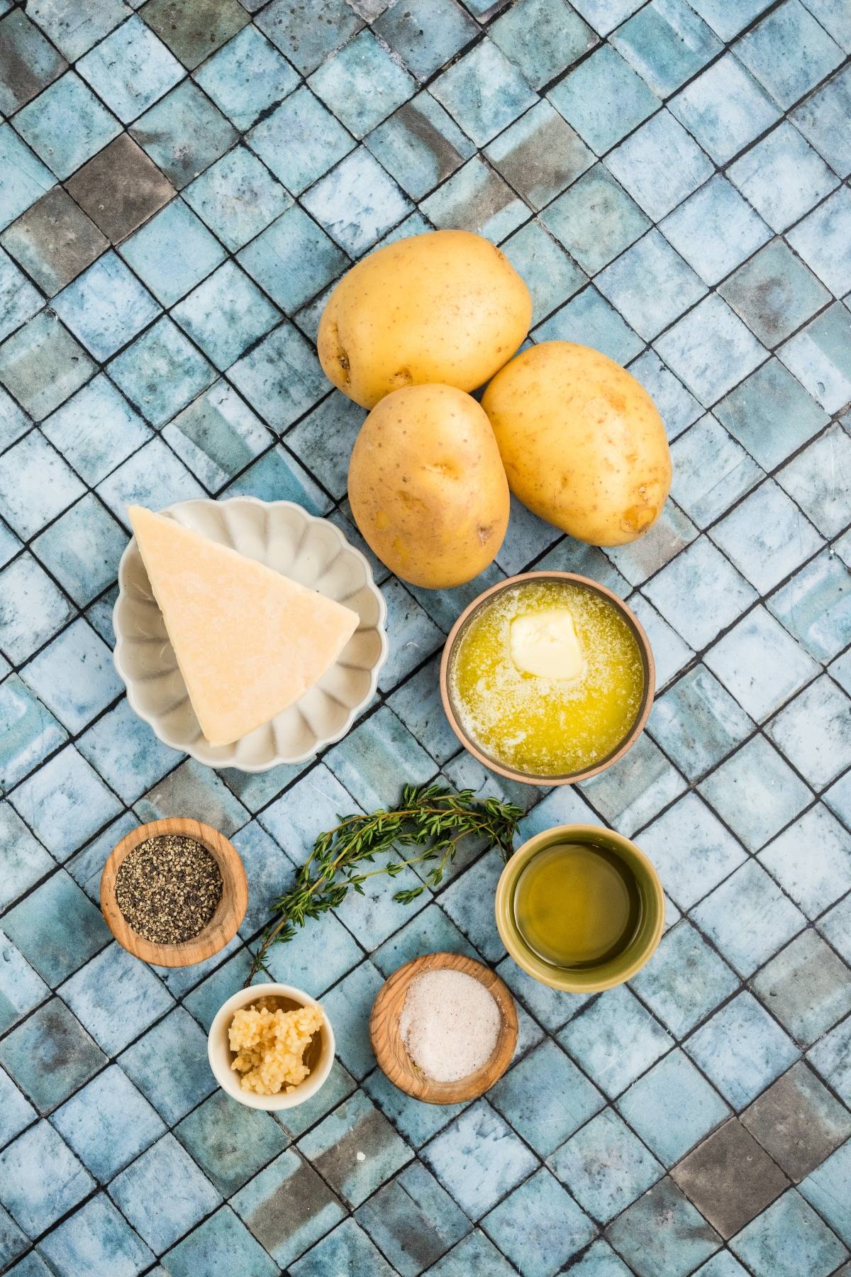 Three potatoes, Parmesan cheese, melted butter, olive oil, garlic, salt, pepper, and thyme on a tiled surface.