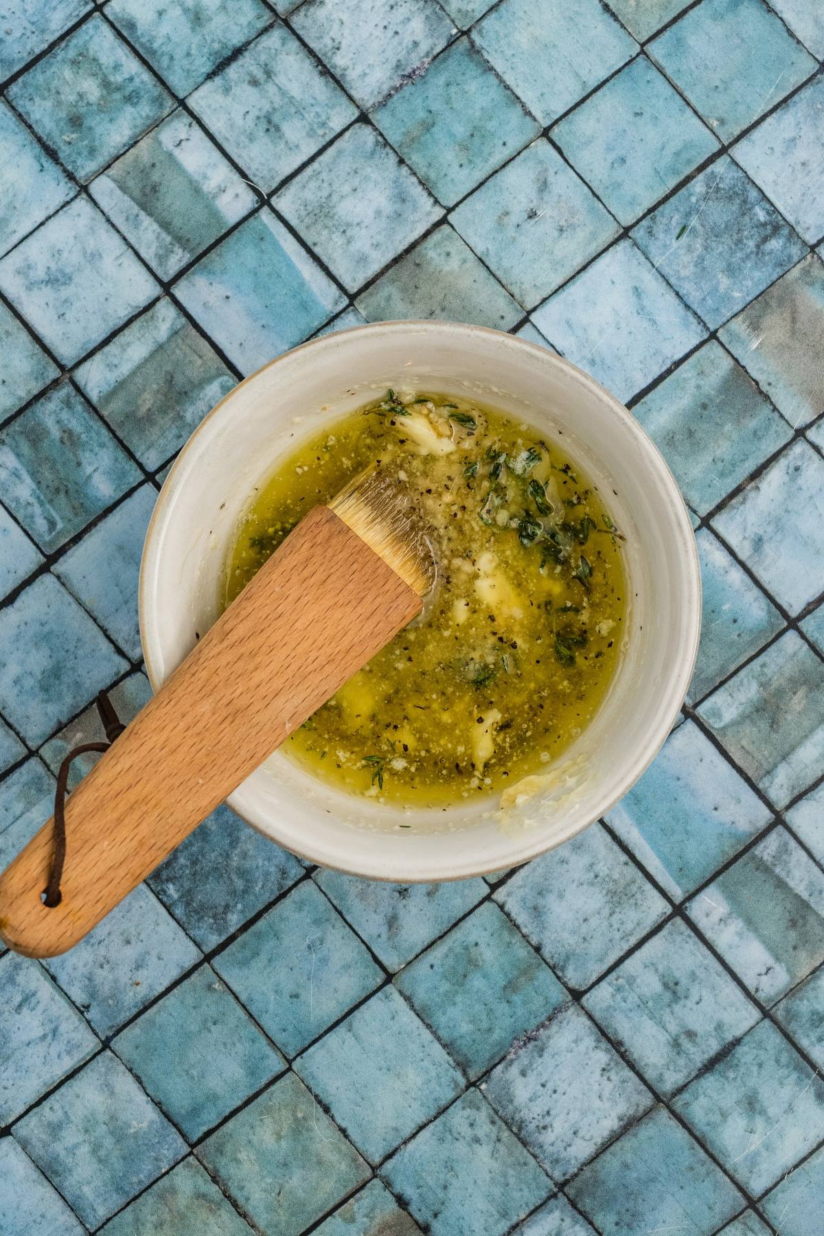 A bowl of herb butter with a wooden brush on a blue tiled surface.