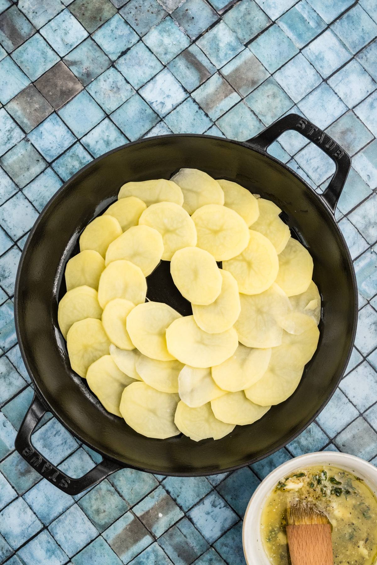 Sliced potatoes arranged in a circular pattern in a black pan on a blue tiled surface.