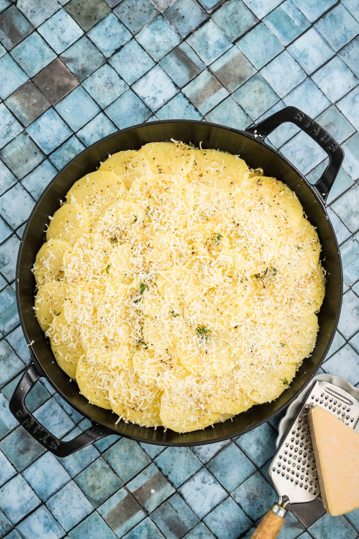 A round baking dish with sliced potatoes topped with grated cheese, next to a cheese grater and cheese wedge.