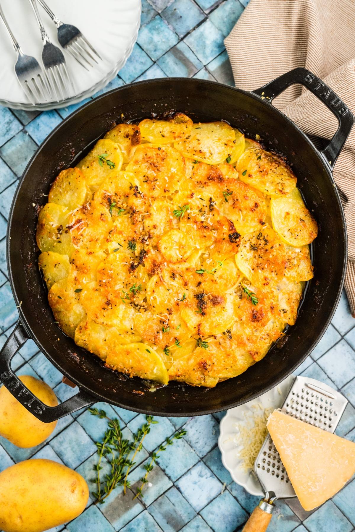 Golden potato galette in a black pan, topped with herbs, next to cheese, grater, and fresh potatoes.