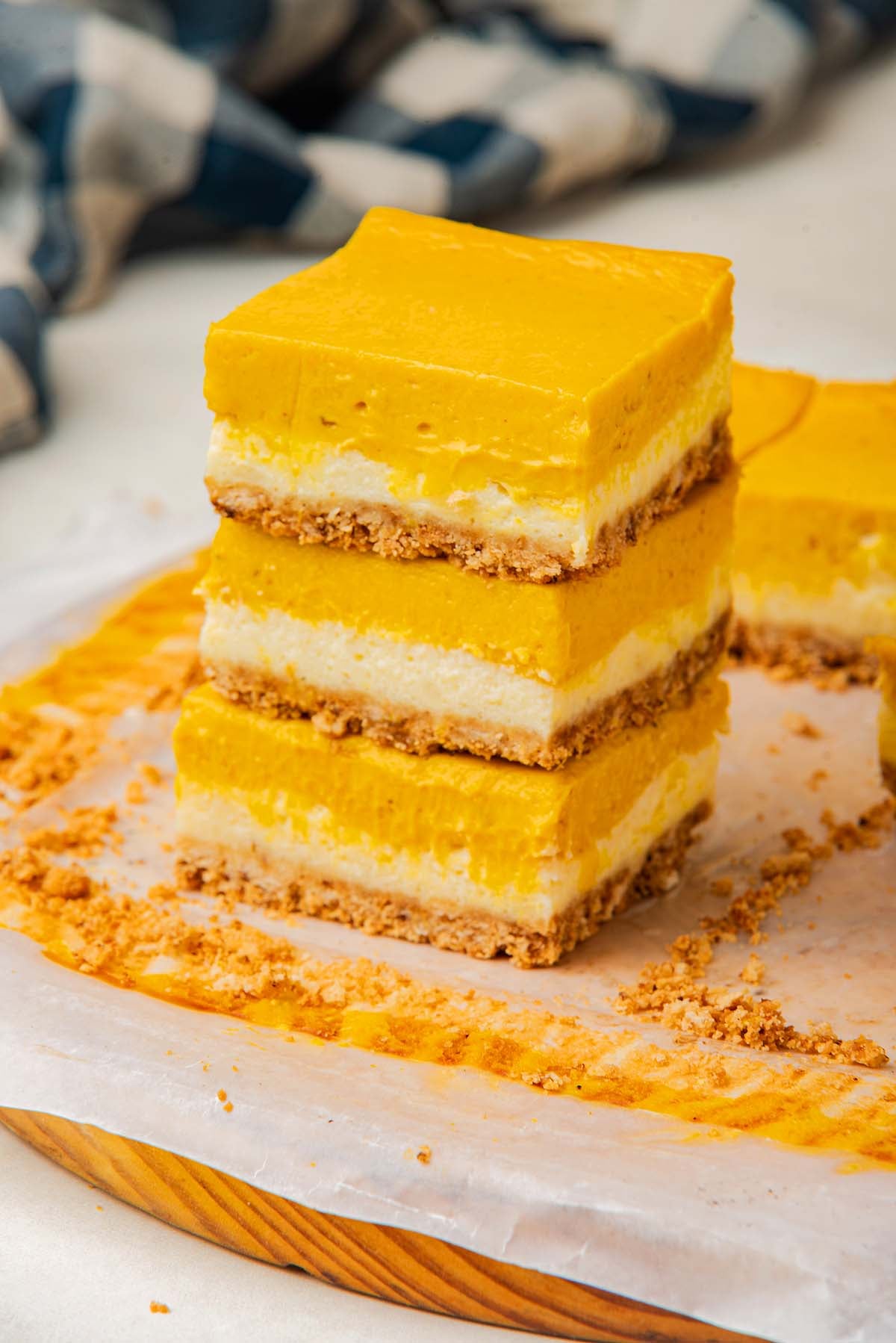 Three pumpkin cheesecake bars stacked on a parchment-lined wooden board, with crumbs scattered around.