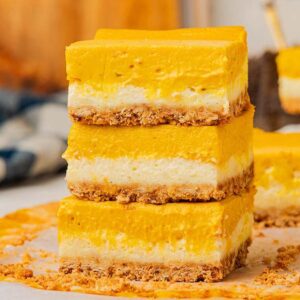 Three stacked slices of pumpkin cheesecake bars with a crumbly crust rest on parchment paper, showcasing their delicious layers.