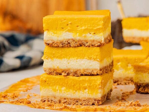 Three stacked slices of pumpkin cheesecake bars with a crumbly crust rest on parchment paper, showcasing their delicious layers.