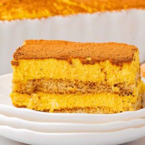A slice of pumpkin tiramisu on a white plate, with more pumpkin tiramisu and ladyfingers in the background.