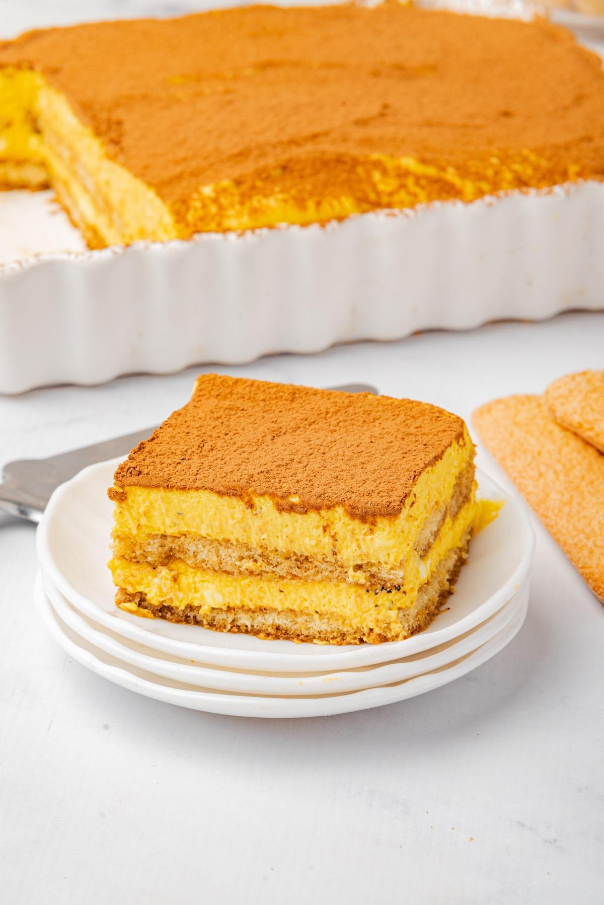 A slice of tiramisu on a plate in front of a larger dish, dusted with cocoa powder.