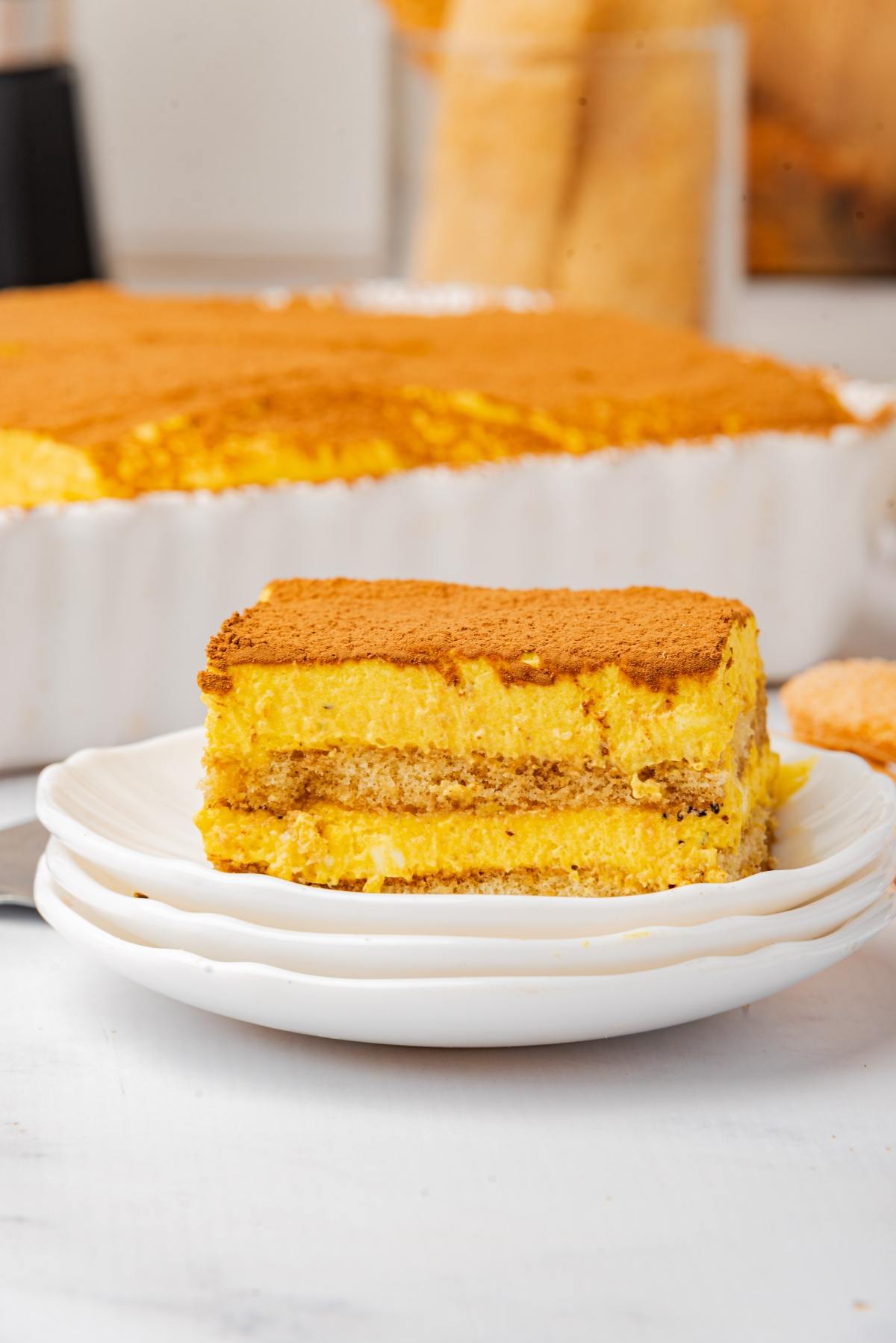 A slice of tiramisu dusted with cocoa powder on a white plate, with more tiramisu in the background.