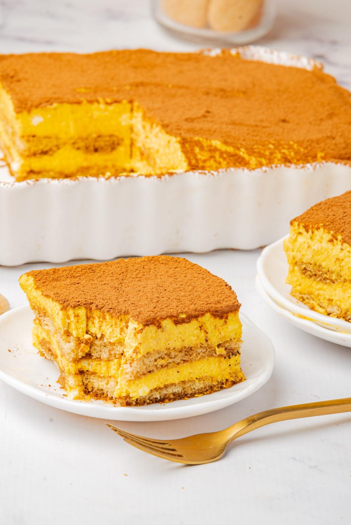A slice of tiramisu dusted with cocoa powder on a plate, with a larger dish of tiramisu in the background.