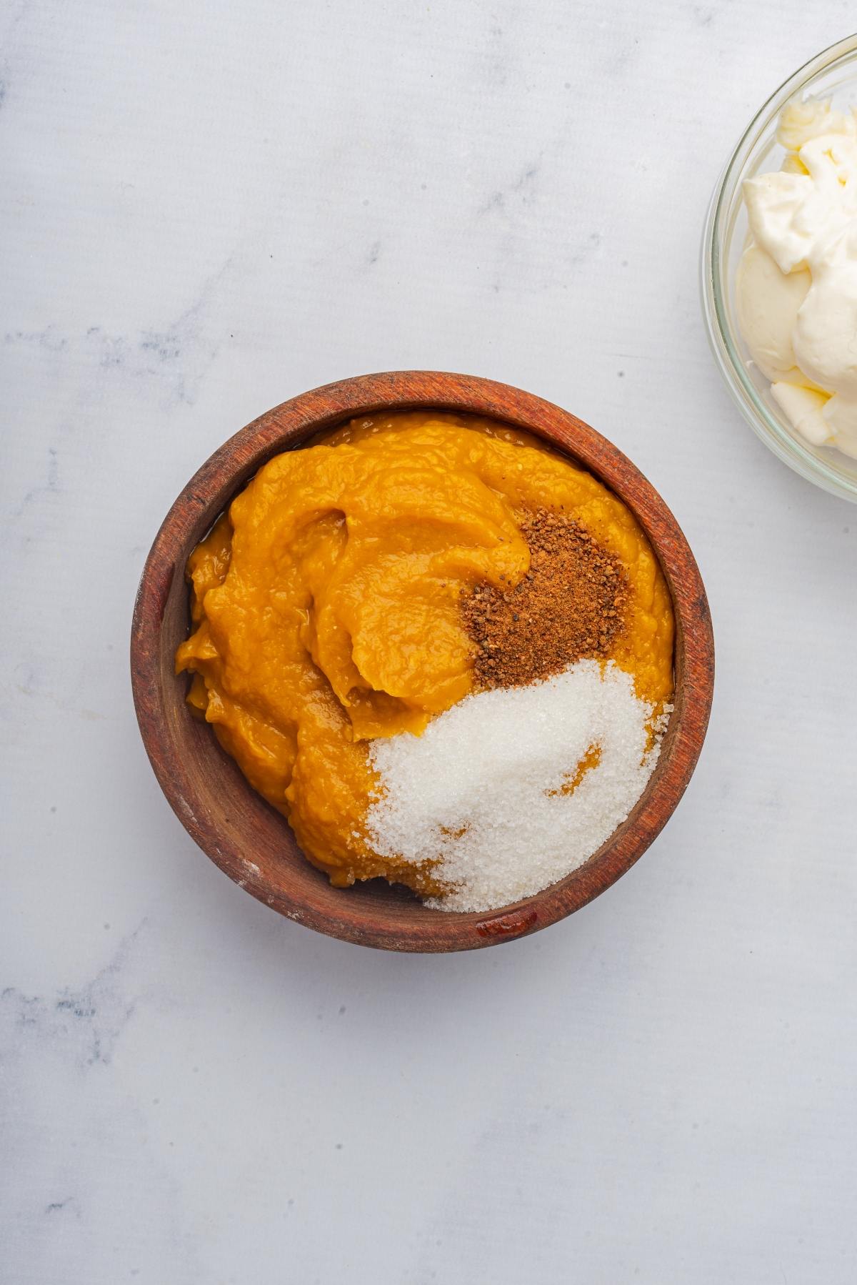 Wooden bowl with pumpkin puree, sugar, and spices next to a glass bowl of cream on a white surface.