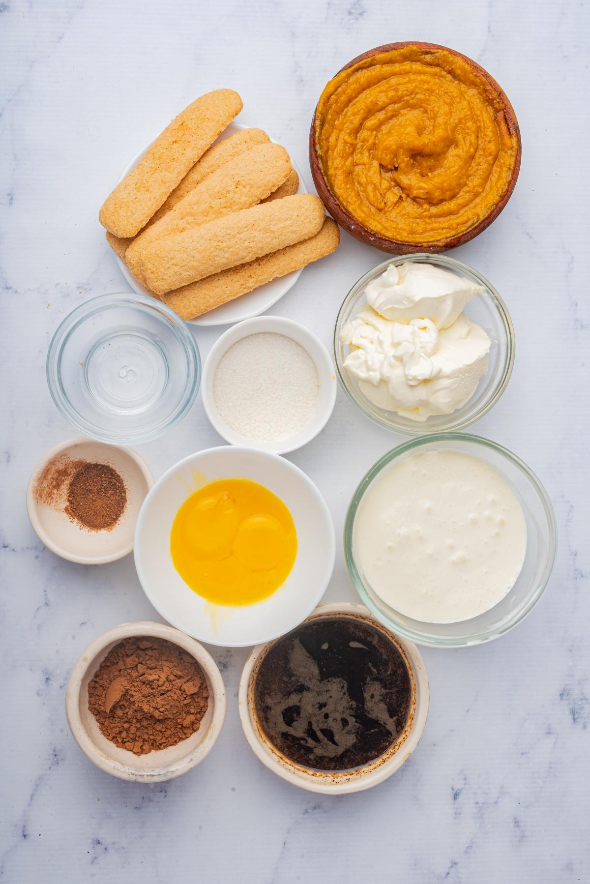 Assorted ingredients for a dessert, including ladyfingers, pumpkin puree, eggs, cream, and spices on a white surface.