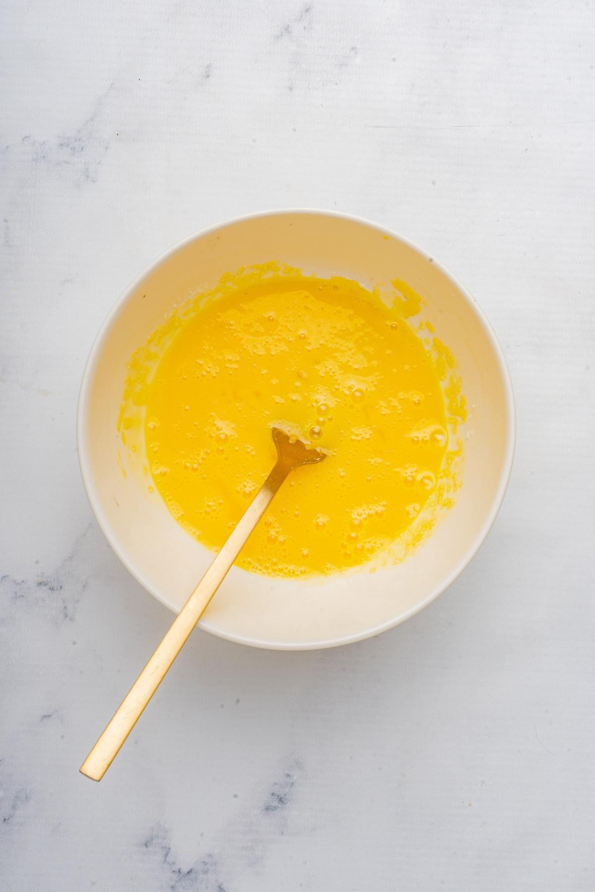 A bowl of beaten eggs with a fork on a light marble surface.